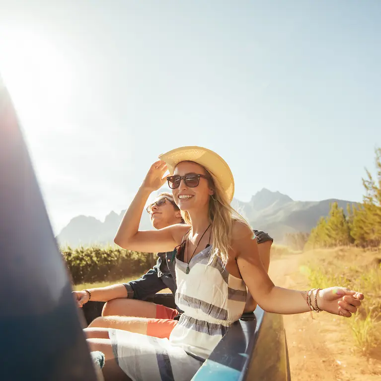 Eine Frau und ein Mann sitzen auf der Ladefläche eines Autos und genießen die Sonne. Im Hintergrund sind die Berge. 