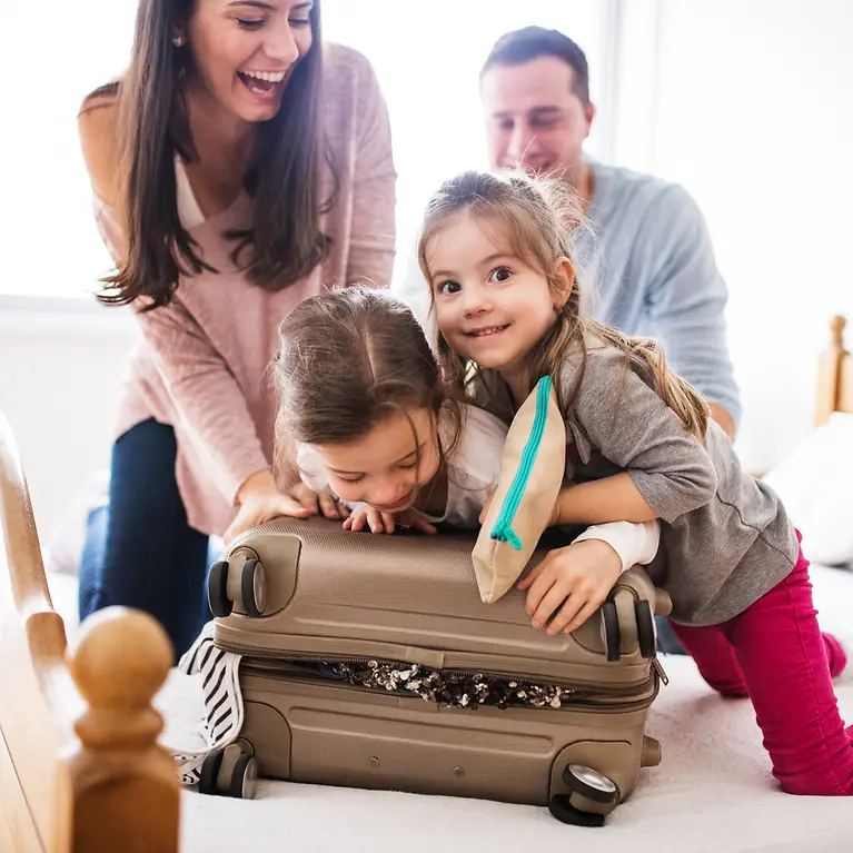 Mutter, Vater und deren zwei Töchter sitzen auf einem Bett und probieren ihren Koffer zu zu bekommen.