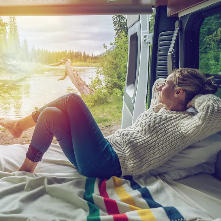 Eine Frau liegt in einem Camper. Im Hintergrund ist ein See zu sehen. 