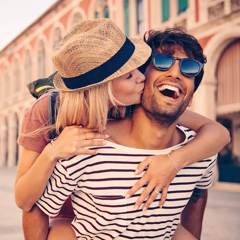 Eine Frau und ein Mann sind in einer Stadt. Er nimmt sie Huckepack und sie gibt ihm einen Kuss auf die Wange. 